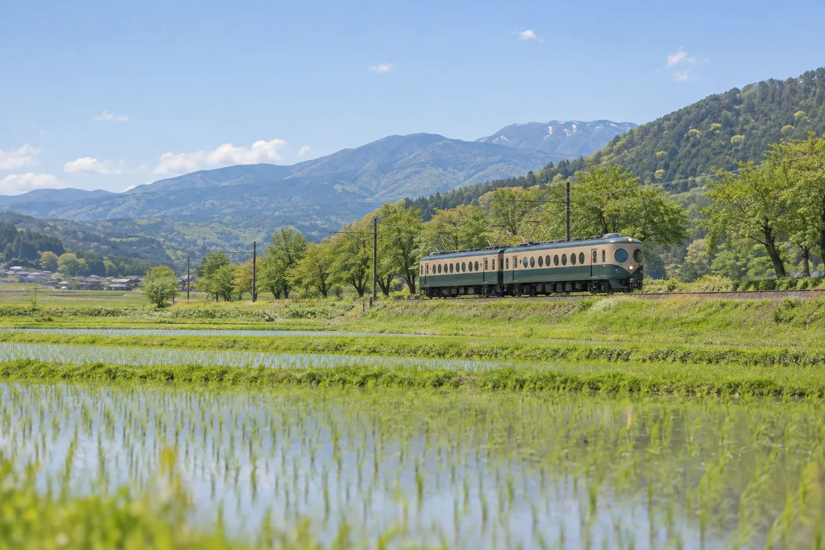 新緑の田園を走る丸窓の古い電車