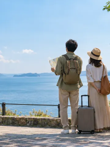 海辺で旅行かばんと並ぶ新婚夫婦の後ろ姿